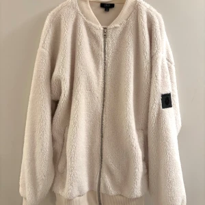Vit teddyjacka från SMOG - Mysig vit teddyjacka från SMOG i relaxed fit. Jackan har dragkedja framtill, ribbade muddar och en svart patch på ärmen. Perfekt för dig som gillar oversized och chill vibe. Fodrad med mjukt material som håller dig varm under kyliga dagar.