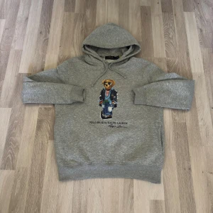 Grå hoodie Polo Bear Ralph Lauren - Säljer en grå hoodie från Polo Ralph Lauren med den ikoniska Polo Bear-trycket framtill. Tröjan har huva med snörning och är långärmad. Perfekt för dig som gillar streetstyle och vill ha något bekvämt men ändå snyggt.