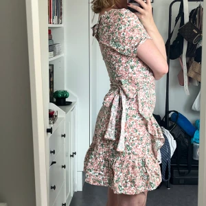 Blommig klänning  - Rosa och grön blommig klänning med små bin på. Den är oanvänd och lappen samt extra knappen är kvar. Den knyts i midjan för att kjolen ska få en omlott-känsla men hela klänningen knyts inte.