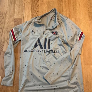 PSG långärmad träningströja Jordan M - Snygg långärmad PSG fotbollströja från Jordan i ljusgrått med svarta stänk och mörkblå logga. Tröjan har dragkedja vid halsen, Paris Saint-Germain-emblem på bröstet och röda/blå detaljer på ärmarna. Tillverkad i funktionsmaterial, perfekt för träning.