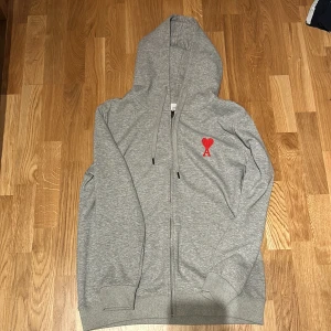 Ami hoodie - Säljer en grå hoodie från Ami Alexandre Mattiussi med dragkedja och huva. Tröjan har en broderad röd hjärtlogga med bokstaven A på bröstet, ribbade muddar och dragsko i huvan. Perfekt för chill dagar och snygg streetstyle. Pris kan diskuteras!