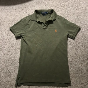 Olivgrön Polo Ralph Lauren piké - Säljer en olivgrön pikétröja från Polo Ralph Lauren i slim fit. Klassisk krage, korta ärmar och orange broderad logga på bröstet. Tillverkad i bomull för en skön känsla och snygg passform. Perfekt för dig som gillar stilrena och tidlösa plagg.