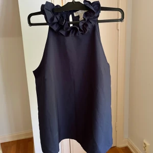 Marinblå ärmlös topp med volangkrage - Snygg marinblå topp från Forever 21 med volangdetalj runt halsen. Modellen är ärmlös och har en loose passform som ger en chill vibe. Perfekt att styla med jeans eller kjol för en trendig look.