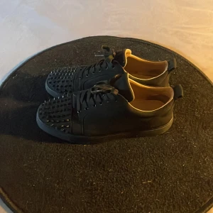 Christian Louboutin skor - Svarta sneakers med coola svarta nitar på tån och klassisk röd sula. Skorna har snörning och är tillverkade i skinn med beige insida. Perfekt för dig som vill sticka ut med en edgy stil. 