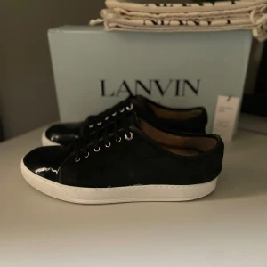 Svarta sneakers från Lanvin - Snygga svarta sneakers från Lanvin med vit platt sula och blanka detaljer på tån. Skorna har snörning med svarta skosnören och metallöljetter. Utsidan är i skinn och insidan är fodrad i ljust läder. Perfekta för dig som gillar stilrena och exklusiva sneakers. 8/10 skick, kritvit sula fixade med hjälp av vit skokräm. Helt nya skosnören på, endast lite smutsig mocka. Storlek UK9 motsvarar 43/44. Original box, två st dustbags och tre st använda skosnören medföljer vid köp (ÄKTIGHETSBEVIS FINNS)