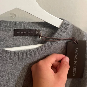 Grå rundhalsad tröja Oscar Jacobson - Klassisk grå rundhalsad tröja från Oscar Jacobson i modellen heter Valter Roundneck. Tröjan är stickad med ribbade muddar vid hals, ärmslut och nederkant. Perfekt inför hösten, lager-på-lager och enkel att matcha med olika outfits! Tröjan är aldrig använd då den är förstor. Nypris: 1499kr