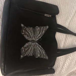 Svart axelväska med strassfjäril från Eternel - Stor svart axelväska från Eternel i syntetmaterial med snygg fjäril i strass på framsidan. Väskan har dubbla handtag, dragkedja upptill och extra dragkedjedetaljer på sidorna. Perfekt för dig som vill ha plats för allt och gillar detaljer som sticker ut.