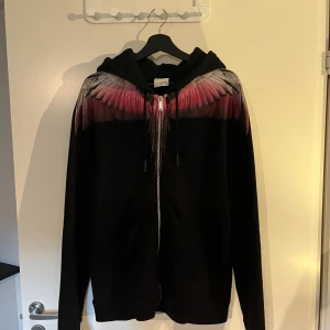 Marcelo Burlon hoddie - Svart hoodie från Marcelo Burlon med dragkedja och huva. Snyggt vingmönster i rött, grått och vitt över axlar och bröst. Långärmad modell med snörning i huvan och två fickor framtill. Perfekt statement-plagg för dig som vill sticka ut.