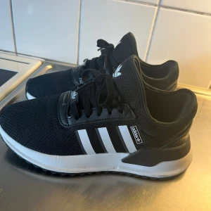 Svarta Adidas sneakers med vita ränder - Svarta Adidas sneakers med klassiska vita ränder på sidorna och vit sula. Ovandelen är i mesh-liknande syntetmaterial och skorna har snörning. Adidas-logga på plösen och sidan. Perfekta för en sportig och stilren look.