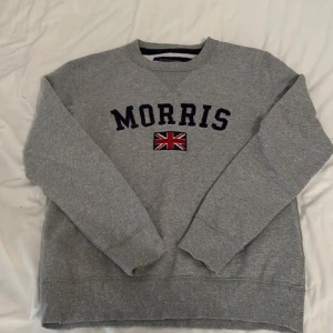 Grå sweatshirt från Morris  - Säljer en grå sweatshirt från Morris i storlek XS. Tröjan har broderad Morris-logga och brittisk flagga på bröstet samt en patch på ärmen. Klassisk rund halsringning och ribbade muddar. Perfekt för en chill och stilren look. Passar XS-S