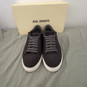 Mörkgrå sneakers från Axel Arigato - Snygga mörkgrå sneakers från Axel Arigato med ovandel i mocka och klassisk vit sula. Skorna har rund tå, platt sula och grå snörning. Perfekta för dig som gillar stilrena och minimalistiska skor med premiumkänsla. Finns även med arigato påsen