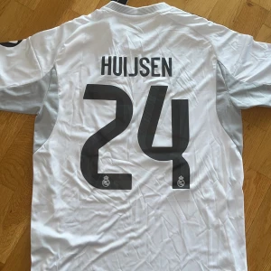 Real Madrid Huijsen #24 fotbollströja S - Säljer en officiell Real Madrid fotbollströja från Adidas i storlek S, med Huijsen och nummer 24 på ryggen. Tröjan är vit med svarta detaljer, gula linjer och klubbmärke på bröstet. Kortärmad och tillverkad i lätt, ventilerande polyester. Slim fit-modell.