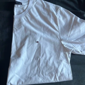 Vit t-shirt från Tommy Hilfiger - Klassisk vit t-shirt från Tommy Hilfiger med rund hals och liten broderad logga på bröstet. Tillverkad i mjuk bomull och har en enkel, stilren design som passar perfekt till jeans eller shorts.