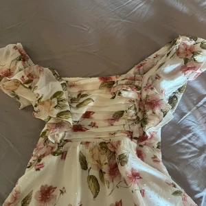 Blommig  blus med volang - Säljer en romantisk blommig blus i vit ton med rosa och gröna blommor. Toppen har volang och en smockad rygg för extra komfort. Materialet är lätt och luftigt, perfekt för varma dagar. Passar dig som gillar en feminin och trendig stil.