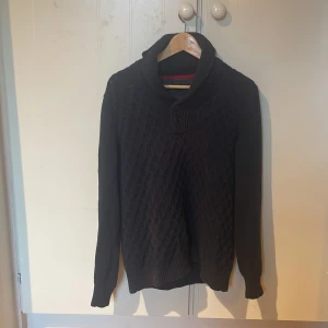 Svart stickad tröja Tommy Hilfiger - Mörk, stickad tröja från Tommy Hilfiger med snygg struktur och hög krage med knappar. Tröjan är långärmad och har ribbade muddar vid ärmslut och nederkant. Perfekt för kyliga dagar och enkel att matcha med jeans eller chinos. Priset är inte hugget i sten. 