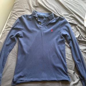Marinblå pikétröja från Ralph Lauren - Snygg marinblå långärmad pikétröja från Polo Ralph Lauren i slim fit. Tröjan har klassisk krage, två knappar vid halsen och den ikoniska röda logon broderad på bröstet. Perfekt för dig som gillar stilrena och tidlösa plagg.