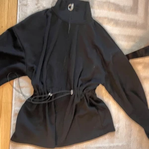 Svart långärmad hoodie med snörning - Svart hoodie med hög krage och dragkedja framtill. Tröjan har snörning i midjan för justerbar passform och långa ärmar. Perfekt för dig som gillar en stilren look med lite edge. Materialet känns mjukt och bekvämt mot huden.