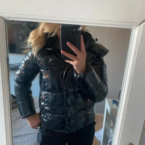 Svart pufferjacka från Calvin Klein - Snygg svart pufferjacka från Calvin Klein Jeans med glansig finish och diskret CK-logo på bröstet. Jackan har huva, dragkedja och är vadderad för extra värme. Perfekt för kalla dagar och ger en cool streetstyle-vibe. Den är kanppt använd och har inga skador alls.