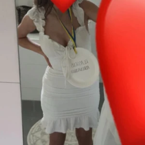 Vit kort volangklänning med knyt - Supersöt vit kort klänning med volangdetaljer på axelbanden och nederkanten. Knytning framtill vid bysten och figurnära passform. Perfekt för sommarens alla tillfällen!