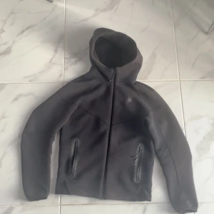 Svart Nike hoodie med dragkedja - Svart hoodie från Nike med hel dragkedja framtill och två fickor med dragkedja. Tröjan har huva och är långärmad, perfekt för kyliga dagar eller när du vill ha en sportig look. Klassisk Nike-logga på bröstet.