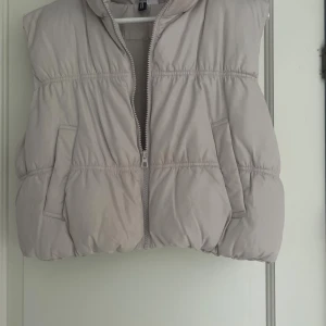 Beige dunväst med dragkedja - Snygg beige dunväst med dragkedja framtill. Västen har två fickor på sidorna och en hög krage som ger extra värme. Perfekt att slänga över en hoodie för en cool och avslappnad look. Det finns även sminkfläckar på kragen.