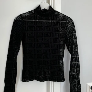 Svart mönstrad polotröja från Amisu - Snygg svart polotröja från Amisu i storlek S. Tröjan har ett genomskinligt meshmaterial med ett grafiskt mönster och lång ärm. Tight passform och hög krage som ger en cool vibe. Perfekt att styla med jeans eller kjol för en trendig look.
