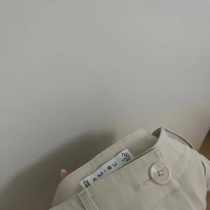 Beiga kostymbyxor från Amisu - Stilrena beiga kostymbyxor från Amisu i storlek 36. Byxorna har en klassisk rak passform med knapp och dragkedja framtill. Perfekta för en clean och snygg look. Materialet känns mjukt och lätt, vilket gör dem bekväma att bära hela dagen.