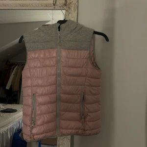 Rosa och grå väst med huva -  dunväst i ljusrosa med grå axelparti och stor huva. Västen har quiltad design, två fickor med dragkedja och hel dragkedja framtill. 