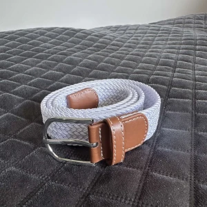 Vitt flätat bälte med bruna skinn-detaljer_ Grisch bälte - Snyggt vitt flätat bälte med detaljer i brunt skinn och silvrigt spänne. Bältet har en stilren look med vit söm längs skinnpartierna och passar perfekt till jeans eller chinos. Flexibelt material som gör det enkelt att justera efter önskad längd.Fungerar for storlekar över 95cm också.