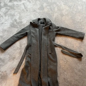 Grå kappa från Bik Bok med bälte XS - Snygg grå kappa från Bik Bok i storlek XS. Kappan har lång passform, bälte i midjan och två fickor framtill. Tillverkad i ullblandning med mjukt foder och klassisk krage. Perfekt för kyligare dagar och ger en clean look.