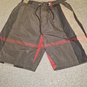 Rutig shorts med snörning - Snygga shorts med rutigt mönster i svart, beige och rött. De har elastisk midja med snörning och praktiska fickor. Perfekta för sommardagar och har en avslappnad passform. Materialet känns lätt och luftigt.