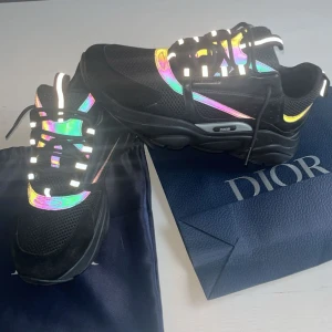Dior sneakers med reflekterande detaljer - Säljer ett par svarta Dior sneakers i storlek 45 som är reflekterande, regnbågsskimrande detaljer. Skorna har mesh och skinnpaneler, samt svarta snören med vita inslag. Kommer med originalpåse och kvitto. Riktigt snygga och unika sneakers för dig som vill sticka ut.