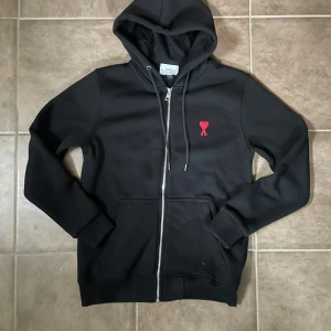 Svart hoodie från Ami med rött hjärta - Svart hoodie från Ami Alexandre Mattiussi med dragkedja, huva och snörning. På bröstet finns ett broderat rött hjärta med A. Tröjan har två fickor fram och är långärmad. Materialet är mjuk bomull, perfekt för chill dagar. Storlek S men sitter som M.