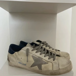 Golden Goose super stars - Golden Goose sneakers i vitt skinn med grå mockadetaljer och ikonisk stjärna på sidan. Skorna har grå snörning, blå hälkappa och platt sula. Perfekta för en avslappnad och trendig look.