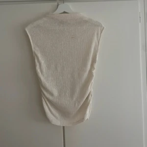 Beige ribbad ärmlös topp - Säljer en beige ärmlös topp med ribbad struktur. Toppen har en enkel och clean design med lätt drapering på sidorna, perfekt för en stilren look. Passar till många olika outfits och är superenkel att matcha.