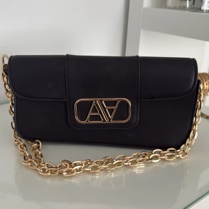 Svart axelväska med guldkedja från AVA - Stilren svart axelväska från AVA i skinn med snygg guldfärgad kedja och logga framtill. Väskan har ett elegant och avlångt format, perfekt för att bära över axeln eller i handen. Guldiga detaljer ger en lyxig känsla och väskan stängs med lock.