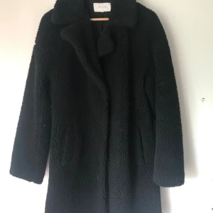 Svart teddyjacka från Vila - Mysig svart teddyjacka från Vila med långärmad design och klassisk krage. Jackan har två fickor framtill och är tillverkad i ett fluffigt teddy-material som ger en cool och trendig look. Perfekt för att hålla värmen under kyliga dagar.
