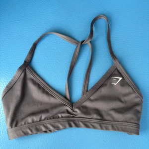 Grå sportig linnetopp från Gymshark XS - Säljer en grå linnetopp från Gymshark i storlek XS. Toppen har smala axelband som korsas i ryggen och en snygg v-ringning framtill. Fler bad finns! Har bara tagit av den för bildens skull. Tillverkad i mjuk och stretchig polyesterblandning, perfekt för träning. Gymshark-logga på bröstet för en sportig vibe. Super snygg grå aktig färg! Använd några få gånger (3-4)