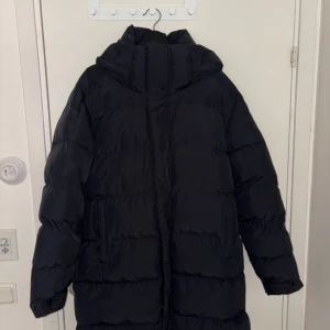 Svart lång pufferjacka med huva ”Everest” - Säljer en svart pufferjacka med huva och dragkedja. Jackan är lång, har två fickor med dragkedja och en riktigt oversized vibe. Perfekt för kalla dagar när man vill vara både varm och stilren.
