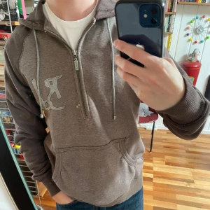 Brun hoodie med half zip - Säljer en brun hoodie från Bondelid med half zip och huva. Tröjan har en stor ficka framtill, snörning i halsen och ett broderat Bondelid-logga på bröstet. Lite mer åt y2k hållet, men passar även till enklare stilar. 