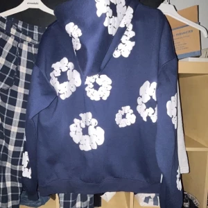 Blå hoodie med vita blommor - Säljer en blå hoodie med stora vita blommönster över hela tröjan. Hoodien har en klassisk huva och en relaxed passform, perfekt för en chill och trendig look. Materialet känns mjukt och skönt mot huden.