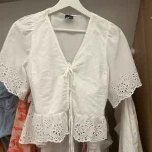 Vit blus med broderade detaljer Gina Tricot - Supersöt vit blus från Gina Tricot i storlek XS. Blusen har v-ringning, knytning framtill och broderade blommiga detaljer på ärmar och nederkant. Kort ärm och volangkant ger en romantisk vibe. Perfekt till sommaren och vårens outfits!