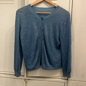 By Malene Birger Glittrig blå kofta - Snygg blå kofta med glittrigt material från By Malene Birger. Koftan har rund hals, långa ärmar och knappar framtill. Tillverkad av viskos, polyester och metalliskt garn som ger en cool shimmer-effekt. Perfekt för dig som vill sticka ut lite extra. Den har lite slitningar men kan lätt fixas med några stygn. Har massor kvar att ge. 