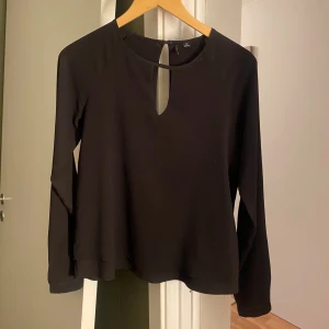 Svart långärmad blus med nyckelhålsdetalj - Säljer en svart långärmad blus  med rund halsringning och snygg nyckelhålsöppning fram och bak. Blusen är i ett lätt, vävt material och har en rak passform som passar till många olika stilar.