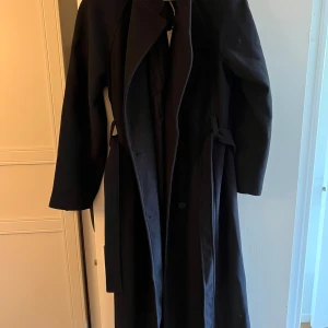 Svart lång kappa från Claire Rose x NAKD - Säljer en svart lång kappa från Claire Rose med axelvaddar och bälte i midjan. Kappan har dubbelknäppning framtill, rak passform och klassisk krage. Materialet är ullblandning som ger en snygg och stilren look. Perfekt för dig som gillar minimalistisk stil. Helt nytt sckick då aldrig använd med etiketten på. 