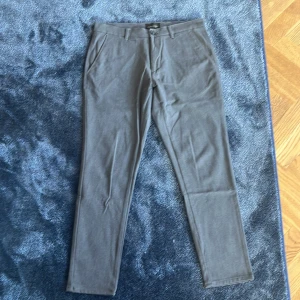 Grå kostymbyxor Tailored Originals - Snygga grå kostymbyxor från Tailored Originals med smal passform. Byxorna har klassiska fickor fram och bak, bälteshällor och stängs med knapp och dragkedja. Perfekta för en stilren look och passar till skjorta eller t-shirt.