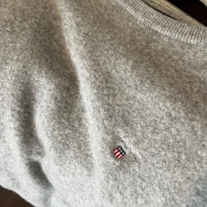 Grå stickad tröja från GANT - Snygg grå stickad tröja från GANT med rund hals och diskret broderad logga på bröstet. Tröjan har ribbade muddar vid ärmslut och nederkant. Materialet är mjukt och värmande ull. Storlek M men liten i storlek.