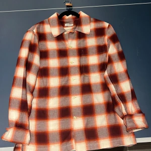 Rutig skjorta från Sandro i orange och vinrött - Säljer en snygg rutig skjorta från Sandro i orange, vinrött och vitt. Skjortan har klassisk krage, knäppning framtill och långa ärmar. Perfekt för dig som gillar statement-plagg med retrovibbar. Bra nu inför hösten som lager på lager samt funkar även som en nize skjorta på sommaren. 