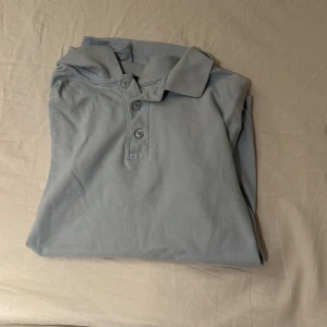 Ljusblå pikétröja från RUN, stl M - Ljusblå pikétröja från RUN i klassisk modell med krage och tre knappar framtill. Tillverkad i bomullstyg som känns mjukt mot huden. Perfekt för en clean och avslappnad stil. Kortärmad och passar bra till jeans eller shorts.