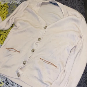 Beige kofta från Ralph Lauren - Klassisk beige kofta från Ralph Lauren med v-ringning och stora knappar framtill. Koftan har två fickor med diskreta detaljer och är tillverkad i ett mjukt, stickat material. Perfekt för lager-på-lager och enkel att matcha med olika outfits.